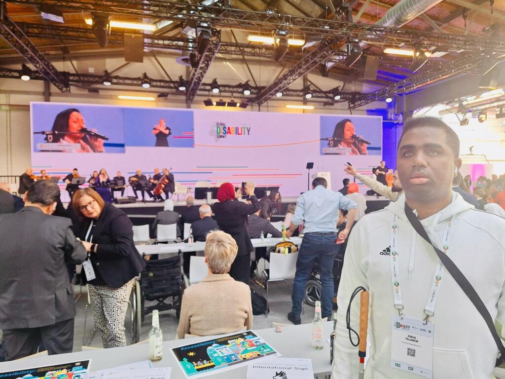 Auf dem Bild ist unser Vorstandsvorsitzender Abbas Yalahow zu sehen. Er steht in einem großen Veranstaltungsraum, trägt einen weißen Adidas-Pullover, ein Namensschild mit der Aufschrift “Abbas Yalahow and Global Disability Summit” und hält einen weißen Stock. Im Hintergrund befinden sich eine Bühne mit Musikern und eine große Leinwand, auf der “Global Disability Summit” sowie eine Frau mit Mikrofon zu sehen sind. Vor der Bühne sitzen zahlreiche Menschen an Tischen mit Broschüren und Flaschen, einige Gäste stehen und unterhalten sich. Die Decke des Raumes ist mit vielen Lichtern und technischen Geräten ausgestattet.
