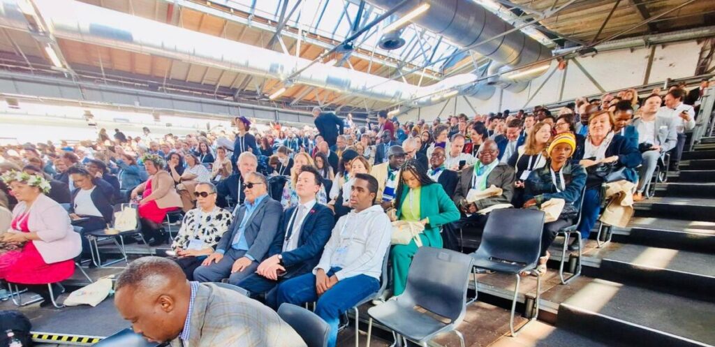 Das Bild zeigt einen großen Veranstaltungsraum, in dem zahlreiche Personen auf Stühlen in geordneten Reihen sitzen. In der ersten Reihe ist unser Vorstandsvorsitzender Abbas Yalahow zu sehen. Der Raum wird von viel Tageslicht durch große Fenster erhellt, und die hohe Decke mit sichtbaren Lüftungsrohren verleiht der Szenerie eine offene Atmosphäre. Die Anwesenden wirken konzentriert und engagiert, während sie an der Veranstaltung oder Konferenz teilnehmen. Einige Gäste sind formell gekleidet, andere erscheinen in legerer Kleidung. Im Vordergrund ist ein Mann in einem karierten Sakko zu erkennen, der auf sein Handy blickt.